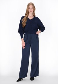 Marineblauwe, textuurgebreide trui met een V-hals en oversized pasvorm, gecombineerd met hoge taille, wijde marineblauwe broeken en zwarte stilettohakken.
