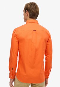 Mężczyzna widziany od tyłu, ubrany w jasnopomarańczową koszulę z długim rękawem i zapiętymi mankietami, oraz beżowe spodnie na tle jednolitego białego tła.