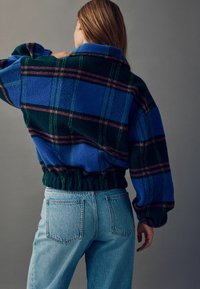 Blau- und grün kariertes Fleece-Pullover mit lockerer Passform, elastischem Bund und Kragen; kombiniert mit hellblauen Jeans.