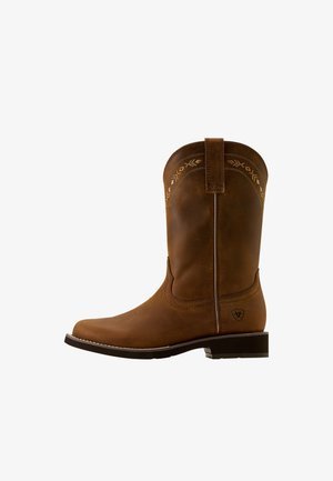 Bota de vaquero de cuero marrón con puntera redondeada, costuras decorativas cerca de la parte superior y un pequeño tacón. Textura lisa y presilla trasera reforzada.