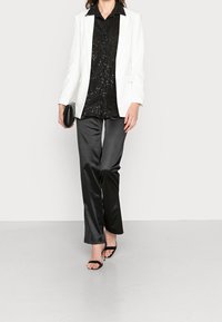 White blazer, black sequined shirt, and black satin pants. Black heeled sandals. Model walking on a light gray floor. Minimalistic background.