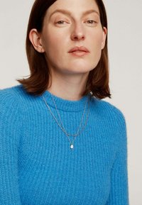 Suéter de punto azul con cuello redondo. Dos collares de capas en tono dorado con un colgante en forma de lágrima. Piel suave y cabello de longitud media.