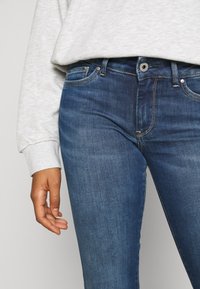 Jeans de mezclilla azul oscuro con un diseño ajustado, sutil efecto de desgaste y costuras en contraste. Combinados con un sudadera de gran tamaño en gris claro.