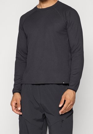 Hombre con camisa negra ajustada de manga larga con textura y pantalones negros con bolsillo con cremallera en el muslo derecho, de pie frente a un fondo liso.