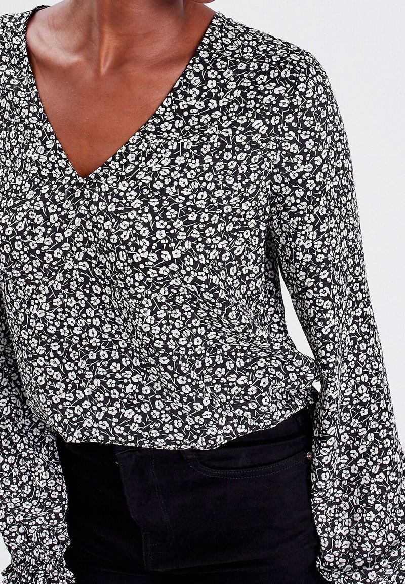 Schwarze Bluse mit weißem Blumenmuster, tiefem V-Ausschnitt und langen, gerafften Ärmeln. Der Stoff wirkt leicht und hat eine glatte Textur.