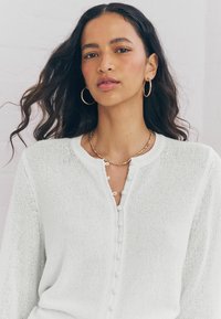 Cardigan en tricot blanc avec texture côtelée, col rond et fermetures à boutons décoratives. Le modèle porte des boucles d'oreilles en or et un collier en perles.