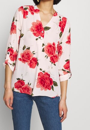 Femme portant un chemisier blanc avec de grands imprimés de roses rouges et des manches trois-quarts, associée à un jean bleu, debout devant un fond uni.