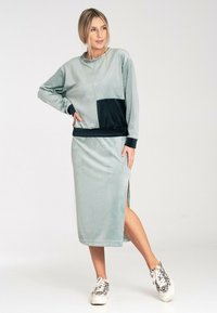 Miękka, jasna zielona sukienka z pluszu z granatową łatką na kieszeni i bocznym rozporkiem, zestawiona ze wzorzystymi trampkami. Luźny krój i długie rękawy.