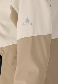 Beige en gebroken wit waterdicht jassenmouw met een klein, grijs, bergvormig logo nabij het bovengedeelte van de arm.