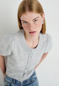 Jonge vrouw met rood haar en sproeten, gekleed in een lichtgrijze button-up trui en blauwe jeans, die recht in de camera kijkt tegen een witte achtergrond.