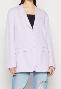 Světle fialový oversized blazer se dvěma knoflíky, klopami a předními kapsami. Nositelný přes černý top a spárovaný se roztrhanými džíny.