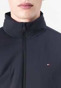 Marineblå jakke med glidelås og høy krage. Laget av lett materiale, har en kompakt glidelås og et lite rødt, hvitt og blått logo.