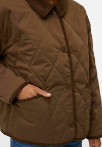 Veste matelassée marron avec un col en velours côtelé, fermeture à boutons et deux poches latérales. Présente des coutures diagonales et un bord contrastant.