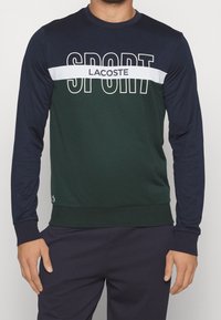 Grön och marinblå långärmad sweatshirt med vit "SPORT" text och "LACOSTE" logotyp. Slätt tyg med ribbad halsringning och ärmslut.