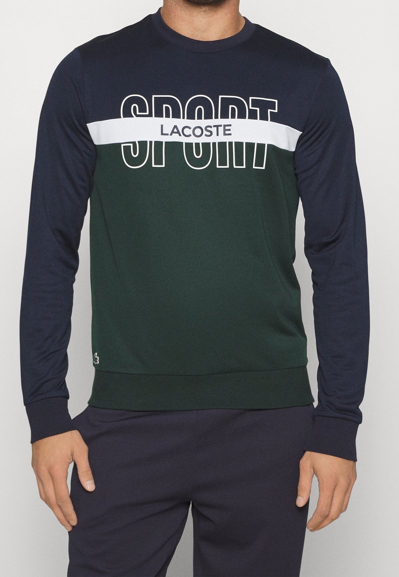 Grön och marinblå långärmad sweatshirt med vit "SPORT" text och "LACOSTE" logotyp. Slätt tyg med ribbad halsringning och ärmslut.