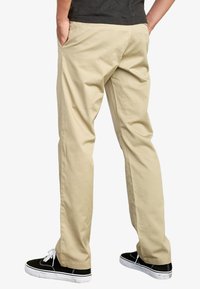 Beige straight-leg pants made from cotton, featuring side pockets and a smooth texture, paired with black sneakers and white laces.