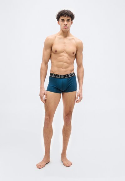 Άνδρας με σγουρά μαλλιά στέκεται ξυπόλυτος, φοράει πετρόλ μποξεράκια με μαύρη ζώνη που φέρει μοτίβο ελληνικού κλειδιού, σε άσπρο φόντο.