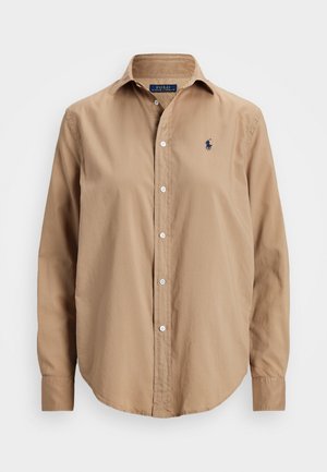 Chemise marron clair à manches longues, boutonnée, avec col et petit petit logo sombre sur la poitrine gauche sur fond blanc.