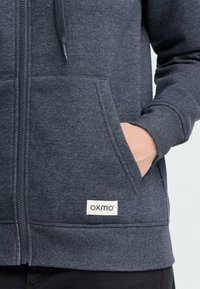 M�ørkegrå zip-up hættetrøje lavet af blødt stof, med en frontlomme og et "oxmo" logo mærke syet på den nederste kant.
