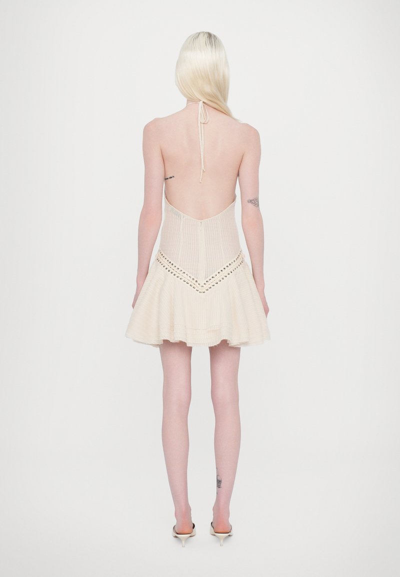 Back view of a sleeveless, light beige dress with a pleated texture, back cutout, and flared skirt design with decorative stitching.