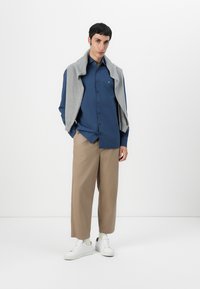 Chemise bleu à boutons, pull gris clair drapé sur les épaules, pantalon large beige et baskets blanches. Design minimaliste et contemporain.