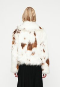 Veste en fausse fourrure avec une base blanche et des taches brunes, dotée d'un col haut, de manches longues et d'une coupe cropped, portée sur une jupe noire.