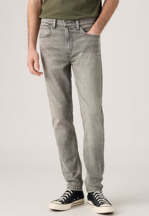 Homme portant un jean gris délavé, des baskets montantes noires et une chemise vert olive, debout avec une main dans la poche.