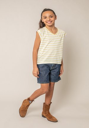 Fille souriante, debout avec une jambe croisée, portant un haut sans manches à rayures jaunes, un short en jean et des bottes en suède marron.