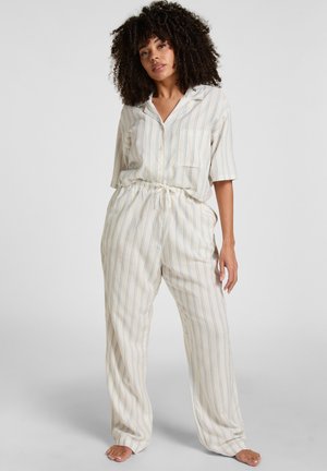 Frau steht barfuß, trägt lockere beige Pyjamas mit dünnen blauen vertikalen Streifen, halbärmliges Hemd mit Knöpfen und Hosen mit Kordelzug.