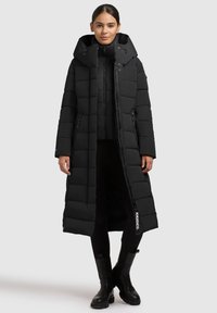 Dlhá čierno-čierna puffová bunda s prešívaným dizajnom, veľkou kapucňou, predným zipsom a bočnými vreckami. Čierne topánky dopĺňajú outfit.