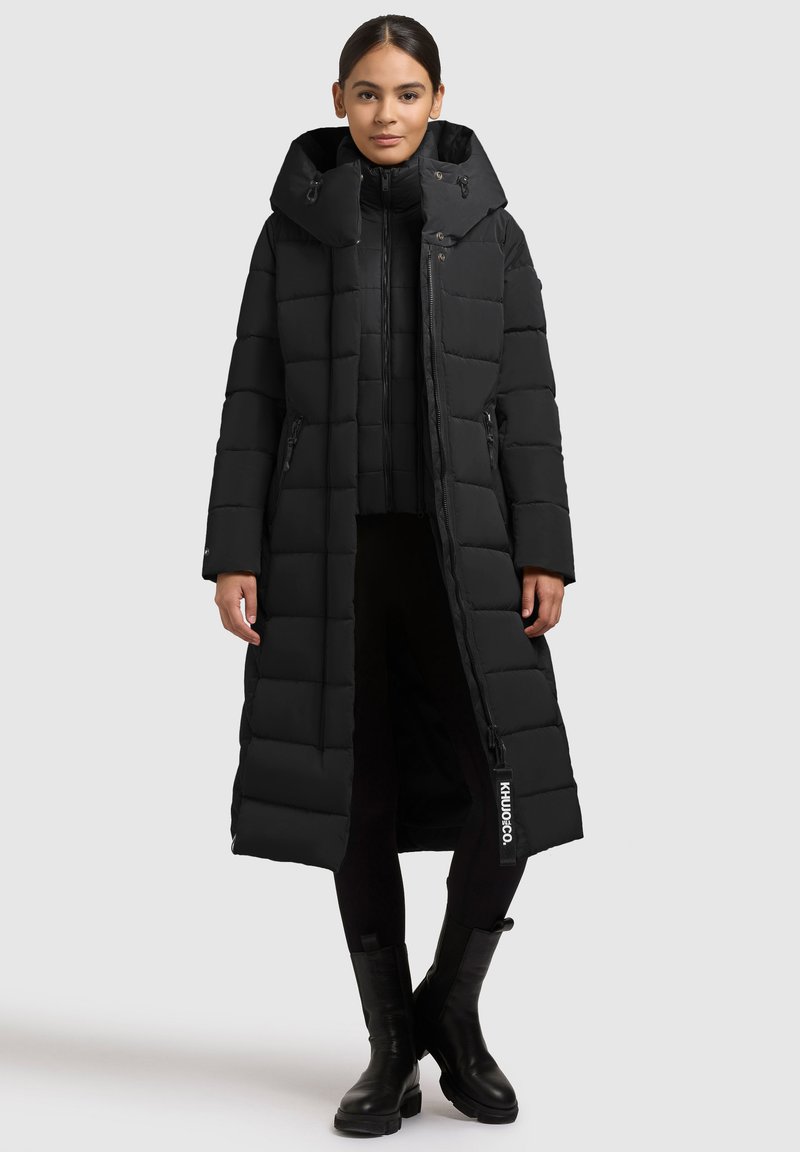 Dlhá čierno-čierna puffová bunda s prešívaným dizajnom, veľkou kapucňou, predným zipsom a bočnými vreckami. Čierne topánky dopĺňajú outfit.