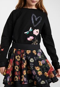 Sweatshirt noir avec broderies florales et d'insectes, associée à une jupe noire évasée ornée de motifs floraux colorés et d'une taille texturée.