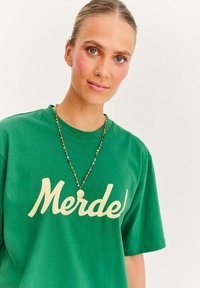 Zelené bavlnené tričko s nápisom "Merdel" v krémovej farbe, doplnené o korálkový náhrdelník s viacerými farebnými korálkami a zlatým príveskom.