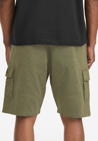 Knee-length cargo shorts in olive green fabric, featuring side pockets, structured stitching, and a relaxed fit. Back view.
