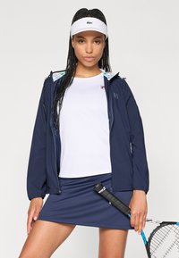 Navy zip-up jacket with light blue accents, white athletic shirt, navy skirt, and holding a tennis racket with a black grip.