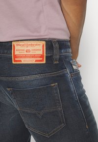 Tmavomodré džínsy z denimu s koženým štítkom, na ktorom sú červené a žlté nápisy, piatimi vreckami a kontrastným prešitím pozdĺž švov.