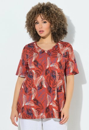 Kvinde med krøllet hår iført en rustfarvet kortærmet top med hvide og marineblå bladmønstre, stående mod en hvid baggrund.