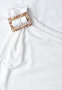 Haut blanc sans manches avec un détail de boucle rectangulaire marron clair rassemblant le tissu sur une épaule.