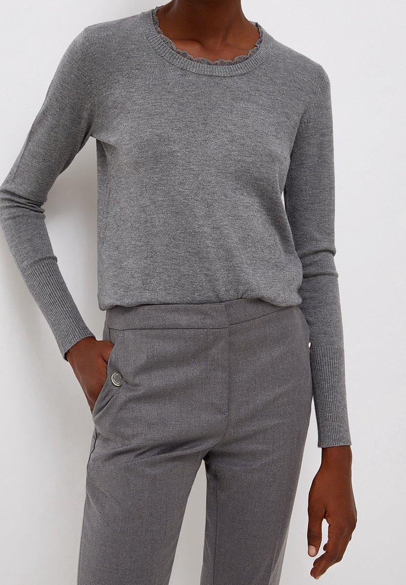 Personne portant un pull gris ajusté à manches longues avec des poignets et un col côtelés, assorti à un pantalon gris taillé sur mesure avec poches latérales.