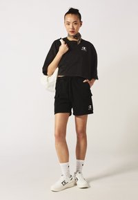 T-shirt nera corta con logo, pantaloni cargo neri, sneakers bianche e calzini alla caviglia bianchi. Include una sciarpa leggera e una borsa a spalla.