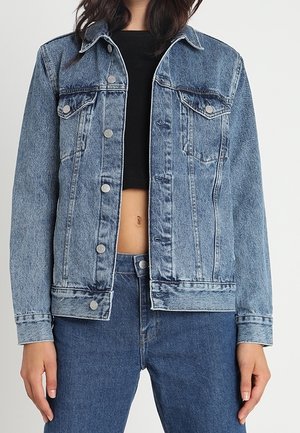 Vrouw draagt een lichtblauwe spijkerjasje over een zwarte cropped top, gecombineerd met bijpassende blauwe spijkerbroek, staand tegen een effen achtergrond.