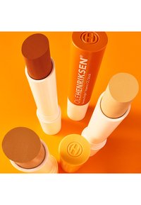Cinque bastoncini cilindrici di CC in diverse sfumature di marrone e beige, con confezioni bianche e arancioni, su uno sfondo arancione brillante.