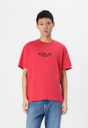 Camiseta de algodón roja con mangas cortas, cuello redondo y logo negro "REPLAY SINCE 1981" impreso en el frente. Llevada con jeans azules.