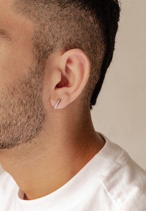 Silberner Creolenohrring am linken Ohr mit einem schlanken, minimalistischen Design. Kurz geschnittenes Haar und strukturierte Haut sind sichtbar.