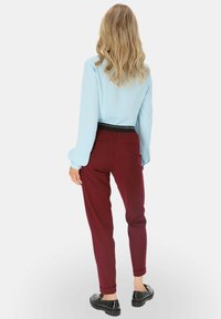 Blondynka z falującymi włosami, ubrana w jasnoniebieską bluzkę, burgundowe spodnie w tapered oraz czarne mokasyny, stojąca na białym tle.