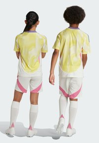 Two kids wearing short-sleeved yellow mesh jerseys with pink accents, white shorts featuring pink stripes, and white socks with pink detailing.
