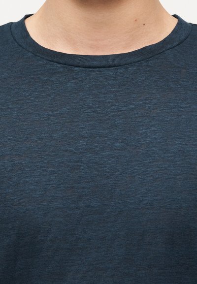 Κοντινό πλάνο του λαιμού και του άνω μέρους του στήθους ενός ατόμου που φοράει σκούρο μπλε μπλουζάκι με στρογγυλή λαιμόκοψη, σε απλό φόντο.