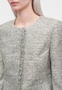 Femme aux longs cheveux clairs portant une veste texturée gris clair avec un col rond et une bordure effilochée à l'avant.