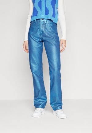 Lyseblå denimjeans med en blank, metallisk finish og rett benkutt. Brukt sammen med en blå mønstret vest og hvite sneakers.