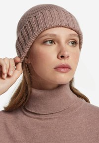 Stickad mössa i mjuk mauve med ribbad struktur; näråliggande design. Modellen bär en matchande högkrage-tröja, vilket visar en minimalistisk stil.